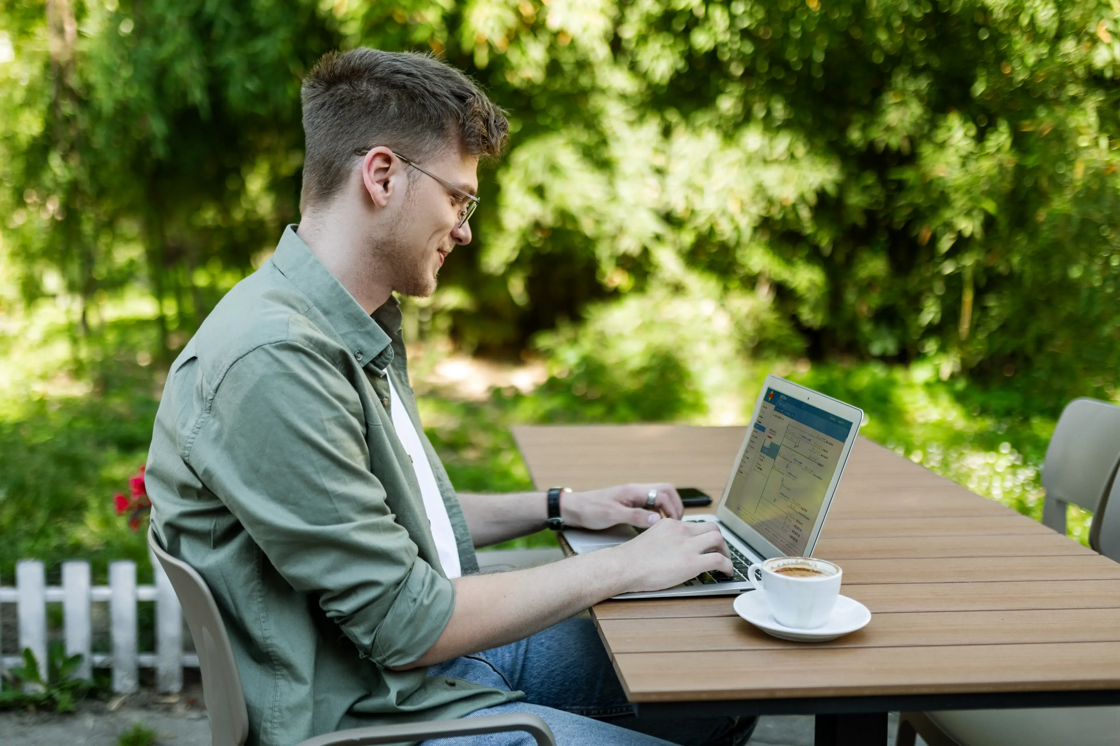 Junger Mann sitzt im Freien an einem Tisch und arbeitet am Laptop mit einer Tasse Kaffee neben sich.